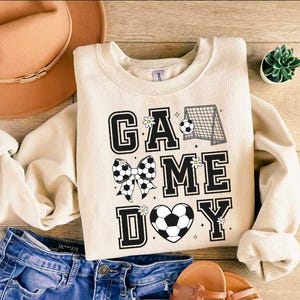 Puede incluir: Una sudadera de color crema con el texto "GAME DAY" en letras negras. Las letras est&aacute;n decoradas con balones de f&uacute;tbol blanco y negro, lazos y flores.