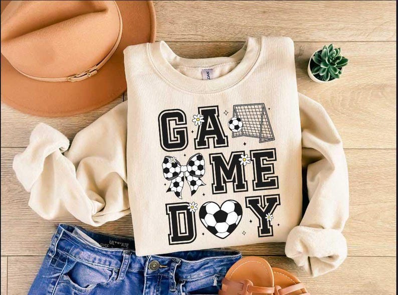 Puede incluir: Una sudadera de color crema con el texto "GAME DAY" en letras negras. Las letras est&aacute;n decoradas con balones de f&uacute;tbol blanco y negro, lazos y flores.