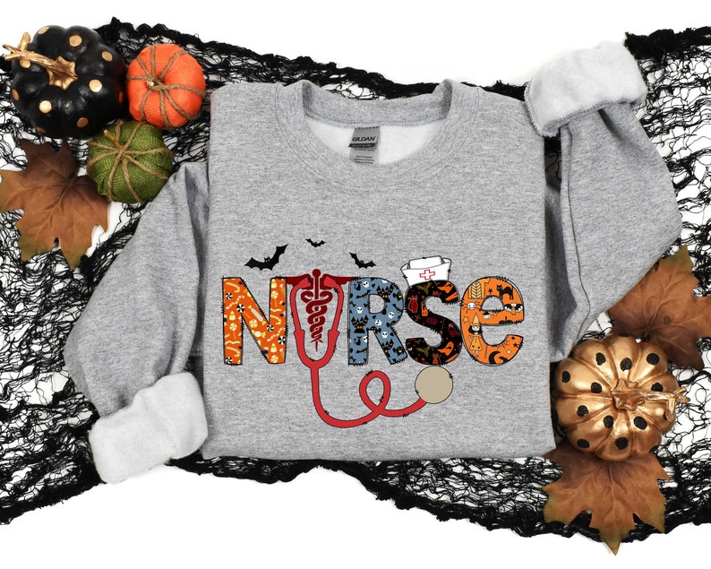 Pu&ograve; includere: Una felpa grigia con un design a tema Halloween che presenta la parola "NURSE" in lettere colorate con uno stetoscopio e pipistrelli.