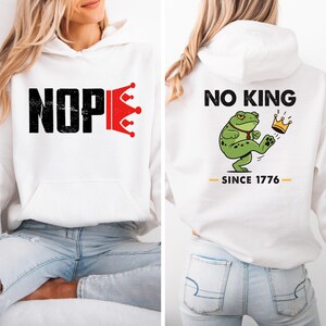May include: White hoodie with the word "NOPE" in black block letters and a red crown graphic. The back of the hoodie features a cartoon frog kicking a crown with the text "NO KING SINCE 1776".