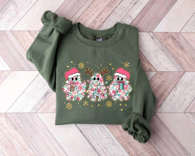 Puede incluir: Una sudadera verde con un dise&ntilde;o festivo que presenta tres fantasmas con gorros de Pap&aacute; Noel y sosteniendo regalos. Los fantasmas est&aacute;n decorados con patrones florales y rodeados de copos de nieve y purpurina.