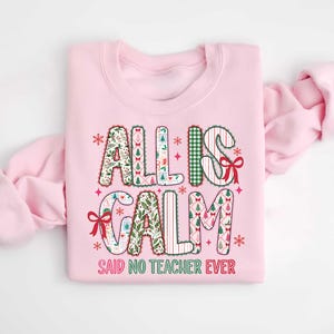 May include: A light pink sweatshirt with the text "ALL IS CALM SAID NO TEACHER EVER". The text is in a festive font with Christmas-themed patterns and red bows. The sweatshirt is folded, with the sleeves draped to the side.