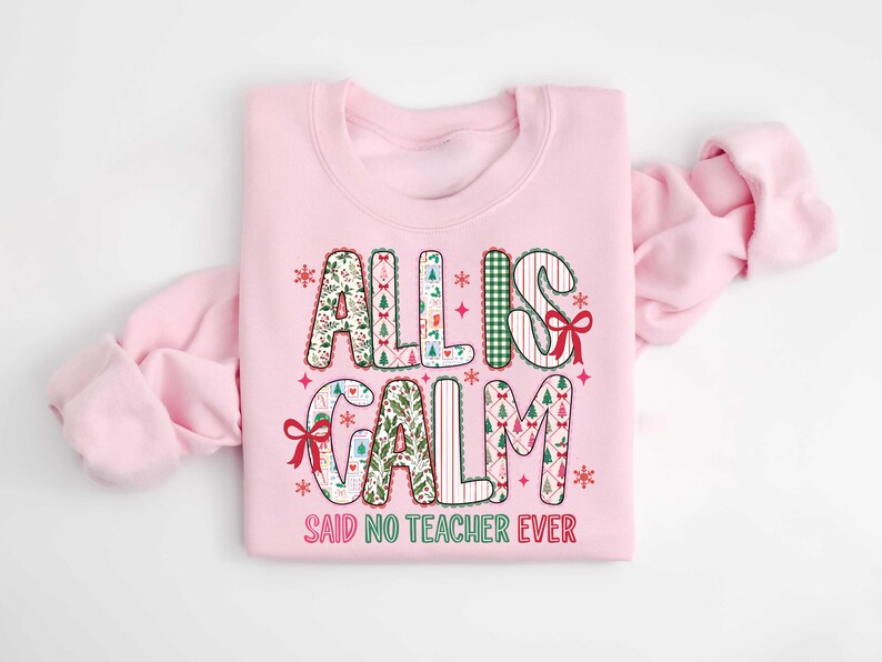 May include: A light pink sweatshirt with the text "ALL IS CALM SAID NO TEACHER EVER". The text is in a festive font with Christmas-themed patterns and red bows. The sweatshirt is folded, with the sleeves draped to the side.