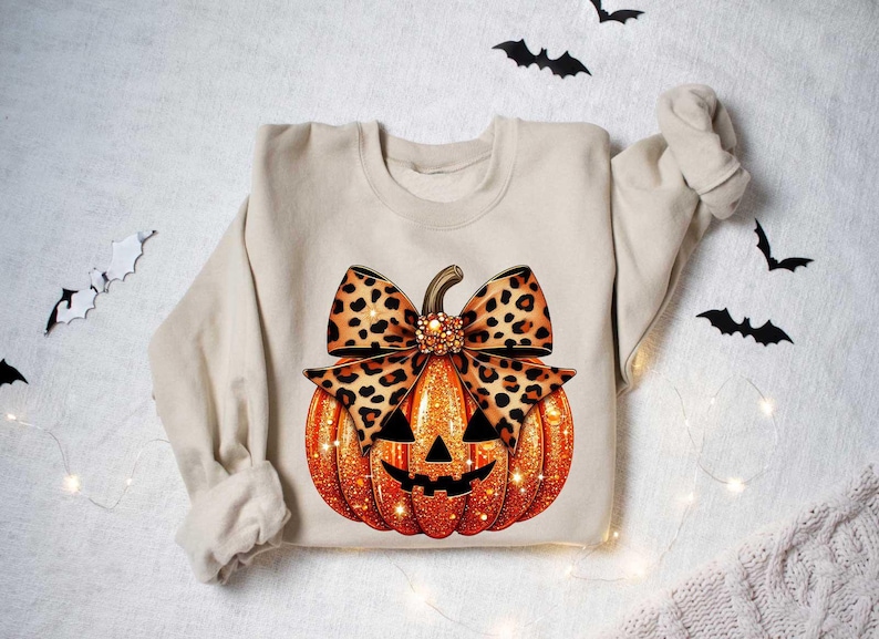 Peut inclure: Un sweat-shirt beige avec une citrouille orange scintillante orn&eacute;e d'un n&oelig;ud &agrave; imprim&eacute; l&eacute;opard. La citrouille a un visage de citrouille d'Halloween.