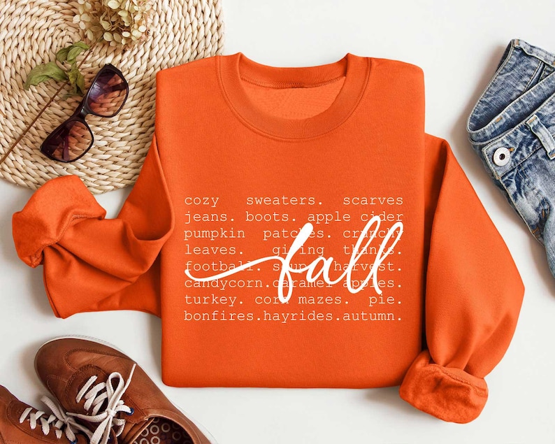 Pu&ograve; includere: Felpa arancione con testo bianco che recita "cozy sweaters, scarves, jeans, boots, apple cider, pumpkin patches, crackling leaves, giving thanks, football, soup, harvest, candy corn, caramel apples, turkey, corn mazes, pie, bonfires, hayrides, autumn." Il testo &egrave; disposto in una colonna verticale sul lato destro della felpa.