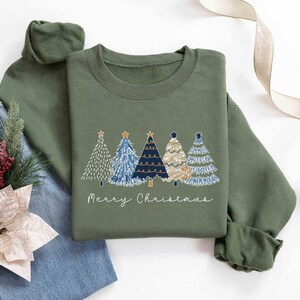 May include: A green sweatshirt featuring a festive design of four stylised Christmas trees in blue, white and gold. The text "Merry Christmas" is printed below the trees.