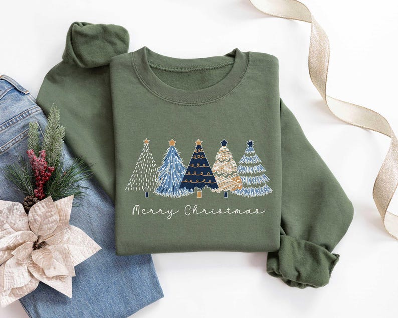 May include: A green sweatshirt featuring a festive design of four stylised Christmas trees in blue, white and gold. The text "Merry Christmas" is printed below the trees.