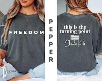 May include: Dark gray t-shirt with the word "FREEDOM" in white on the front. The back of the shirt features the text "this is the turning point" above an American flag graphic and the name "Charlie Kirk".