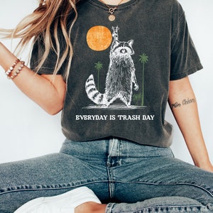 Może przedstawiać: Szary t-shirt z nadrukiem przedstawiającym szopa pracza podnoszącego znak pokoju. Szop stoi przed ż&oacute;łtym słońcem i palmami. Pod szopem znajduje się napis "EVERYDAY IS TRASH DAY".