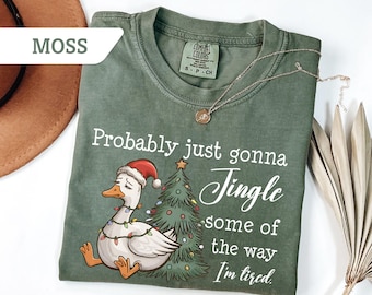 May include: Moss green t-shirt with a Christmas goose graphic and the text "Probably just gonna Jingle some of the way I'm tired." A gold necklace is draped on the shirt. The shirt is made of a soft material.