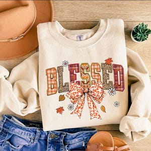 May include: Cream-colored sweatshirt with the word "BLESSED" in a fall-themed design. The letters feature plaid, leopard print, and floral accents. A large bow and autumn leaves complete the design. Blue jeans and a hat are also in the image.