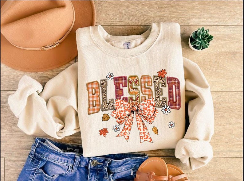 May include: Cream-colored sweatshirt with the word "BLESSED" in a fall-themed design. The letters feature plaid, leopard print, and floral accents. A large bow and autumn leaves complete the design. Blue jeans and a hat are also in the image.