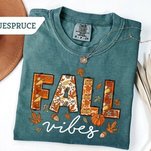 May include: Teal-colored t-shirt with the words "FALL vibes" in a fall-themed design. The word "FALL" is in orange and brown with leaf and acorn details. A gold necklace and a brown hat are also visible.