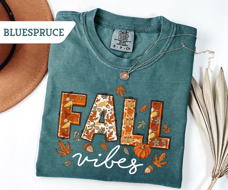 May include: Teal-colored t-shirt with the words "FALL vibes" in a fall-themed design. The word "FALL" is in orange and brown with leaf and acorn details. A gold necklace and a brown hat are also visible.