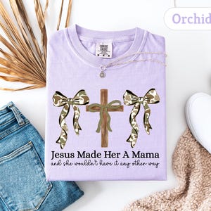 May include: A light purple t-shirt with the text "Jesus Made Her A Mama and she wouldn't have it any other way." The shirt features a cross and two bows with a camouflage pattern. A necklace and white shoes are also visible.