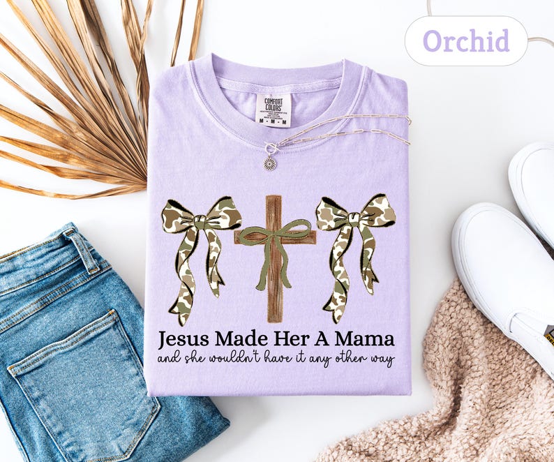 May include: A light purple t-shirt with the text "Jesus Made Her A Mama and she wouldn't have it any other way." The shirt features a cross and two bows with a camouflage pattern. A necklace and white shoes are also visible.