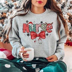 May include: A gray sweatshirt with a Christmas-themed design that says "Nurse" in colorful letters with a red bow and a stethoscope.