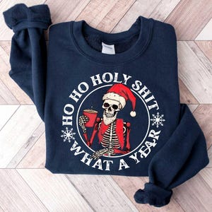 May include: Navy blue sweatshirt with a white graphic of a skeleton wearing a Santa hat and holding a mug. The graphic says "Ho Ho Holy Shit What A Year".