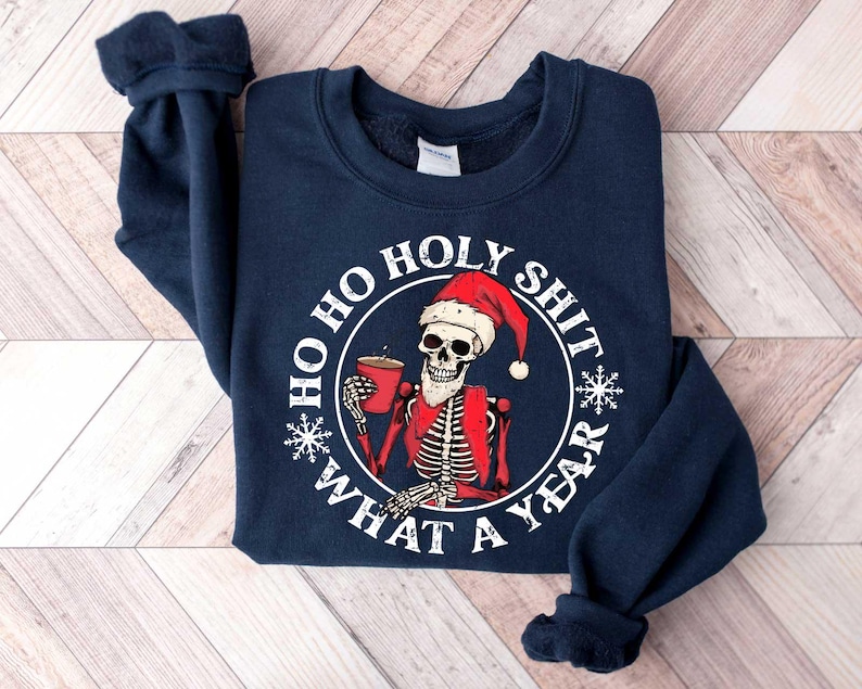 May include: Navy blue sweatshirt with a white graphic of a skeleton wearing a Santa hat and holding a mug. The graphic says "Ho Ho Holy Shit What A Year".
