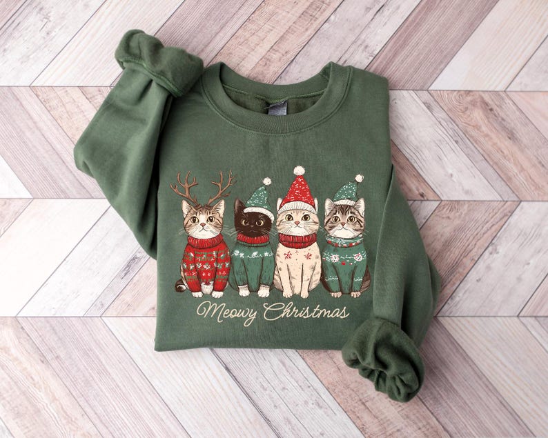May include: Green sweatshirt with a Christmas-themed design featuring four cats wearing Santa hats and sweaters. The text "Meowy Christmas" is printed below the cats.