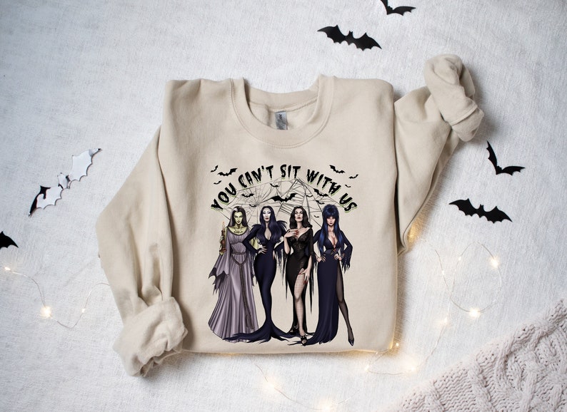 May include: A beige sweatshirt with a graphic print of four women dressed in black and purple gowns. The text "You can't sit with us" is printed in a green circle around the women. The background of the graphic is a black spiderweb with bats flying around it.