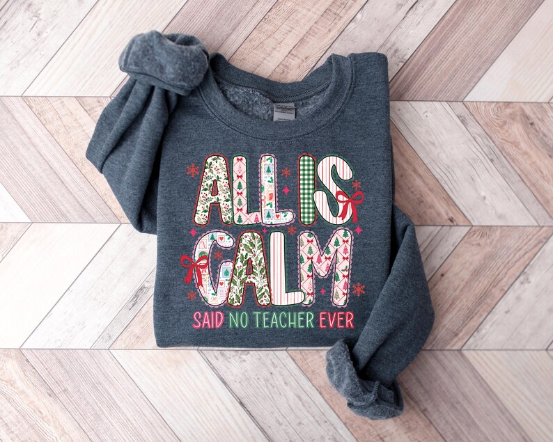 May include: A dark gray sweatshirt with the words "ALL IS CALM SAID NO TEACHER EVER" in a festive, colorful design. The text features Christmas tree and snowflake accents, perfect for the holiday season. The sweatshirt is folded on a wooden surface.