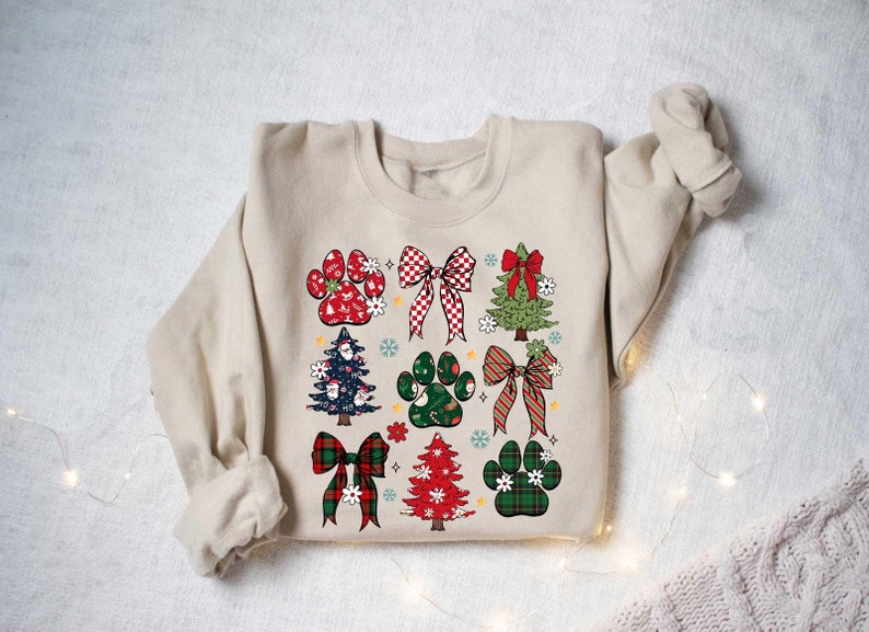 May include: A beige sweatshirt with a Christmas-themed design featuring paw prints, bows, and Christmas trees. The design is in red, green, and white.