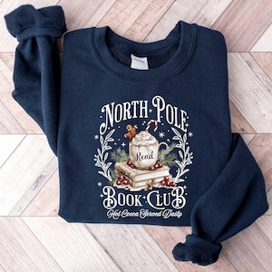 May include: A navy blue sweatshirt with a white graphic design that says "North Pole Book Club" with a mug of hot cocoa, a gingerbread man, and books. The text below the graphic says "Hot Cocoa Served Daily".