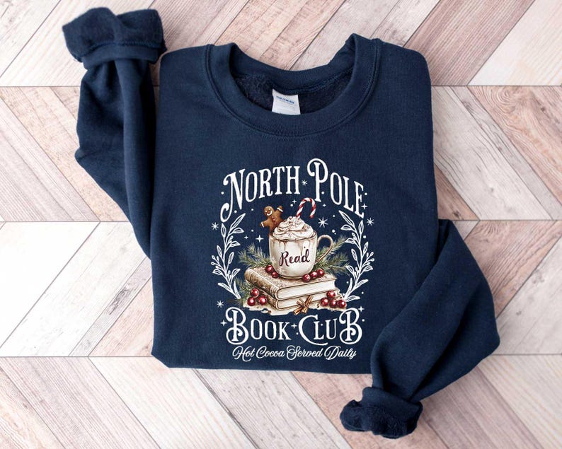 May include: A navy blue sweatshirt with a white graphic design that says "North Pole Book Club" with a mug of hot cocoa, a gingerbread man, and books. The text below the graphic says "Hot Cocoa Served Daily".