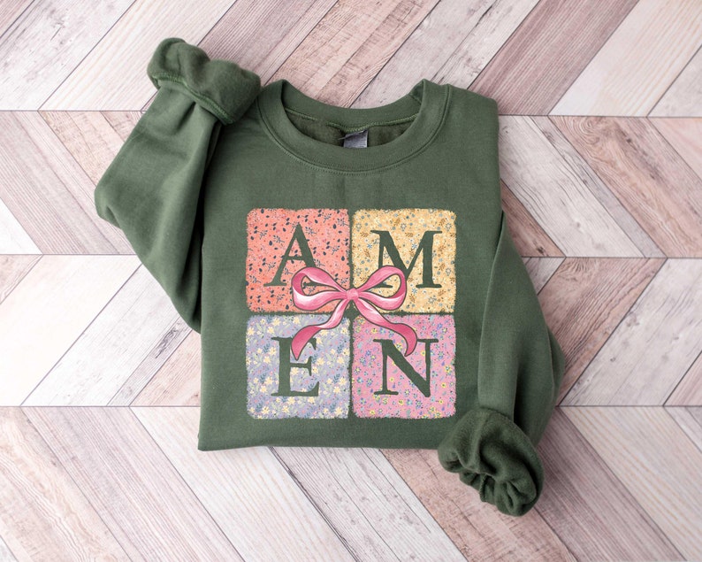K&ouml;nnte beinhalten: Ein gr&uuml;ner Pullover mit einem Aufdruck des Wortes "AMEN" in Patchwork-Optik mit einer rosa Schleife. Die Buchstaben sind von Blumenmustern umgeben.