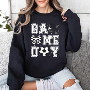 Puede incluir: Sudadera negra con un dise&ntilde;o gr&aacute;fico blanco que dice "Game Day" con un bal&oacute;n de f&uacute;tbol, un lazo y una porter&iacute;a.