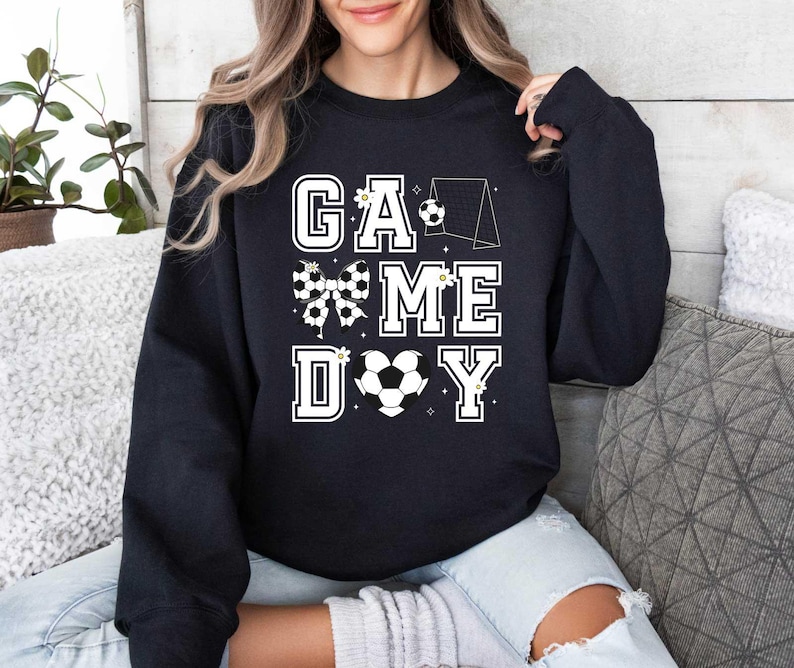 Puede incluir: Sudadera negra con un dise&ntilde;o gr&aacute;fico blanco que dice "Game Day" con un bal&oacute;n de f&uacute;tbol, un lazo y una porter&iacute;a.