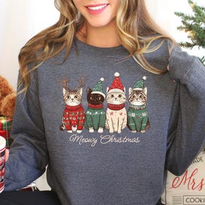 May include: A gray sweatshirt with a Christmas-themed design featuring four cats wearing Santa hats and sweaters. The text "Meowy Christmas" is printed below the cats.