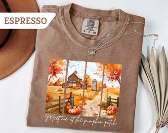 May include: Espresso-colored t-shirt with a fall-themed graphic. The graphic features a barn, pumpkins, and the text "Meet me at the pumpkin patch." A silver necklace with a pendant is draped on the shirt. The shirt is made of a soft material.