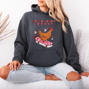 K&ouml;nnte beinhalten: Dunkelgrauer Kapuzenpullover mit einem Aufdruck eines Huhns auf einem Skateboard. Das Huhn tr&auml;gt einen Hut, das Skateboard ist rosa mit Herzen. Der Text "OFF TO COMMIT TOMFOOLERY" steht dar&uuml;ber.