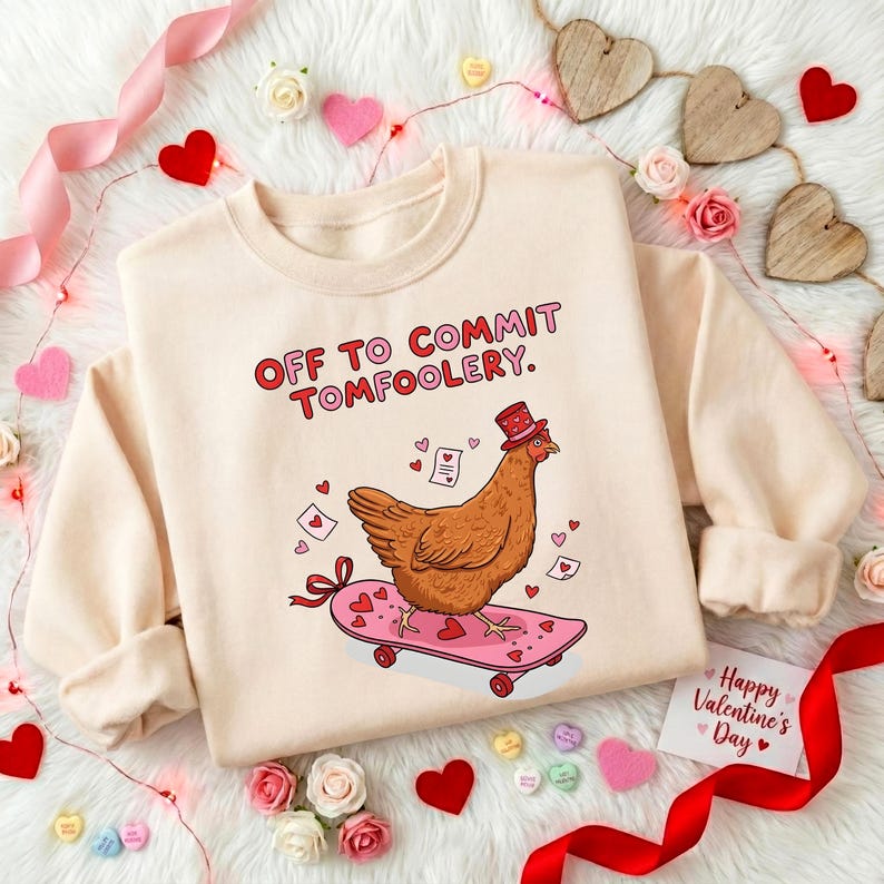 K&ouml;nnte beinhalten: Ein cremefarbenes Sweatshirt mit dem Text "OFF TO COMMIT TOMFOOLERY." Ein Cartoon-Huhn mit Hut f&auml;hrt auf einem rosa Skateboard mit Herzen. Das Design umfasst Herzen und Valentinstagselemente.