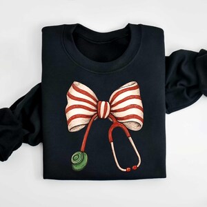 May include: Black sweatshirt featuring a whimsical design of a red and white striped bow and a stethoscope. The stethoscope has red tubing and a green earpiece. The sleeves are black.