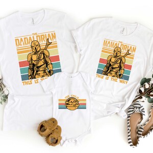 May include: White t-shirts with a retro design featuring a Mandalorian character and the text "The Dadalorian", "The Mamalorian", and "The Child". The shirts have the text "This is the way" printed on them.