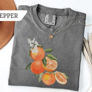 May include: A gray Comfort Colors t-shirt featuring a watercolor illustration of oranges with green leaves and white flowers. A silver necklace with a pendant is on the shirt. The word "PEPPER" is on a white label.