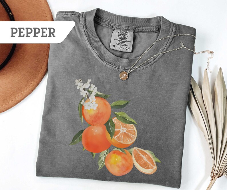 May include: A gray Comfort Colors t-shirt featuring a watercolor illustration of oranges with green leaves and white flowers. A silver necklace with a pendant is on the shirt. The word "PEPPER" is on a white label.