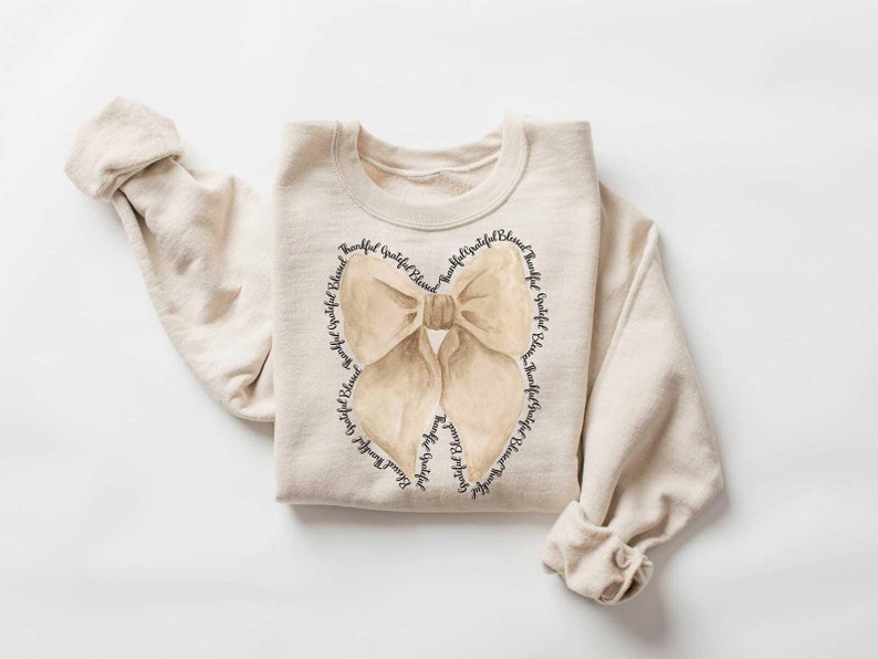 May include: A beige sweatshirt with a brown bow design. The bow is surrounded by the words "Beautiful, Grateful, Blessed" repeated in a circle.