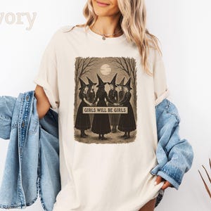 K&ouml;nnte beinhalten: Elfenbeinfarbenes T-Shirt mit einem sepiafarbenen Aufdruck von f&uuml;nf Hexen in schwarzen Kleidern und spitzen H&uuml;ten, die sich im Kreis an den H&auml;nden halten. Der Text "GIRLS WILL BE GIRLS" ist unter der Illustration zu sehen. Das Shirt hat eine lockere Passform.