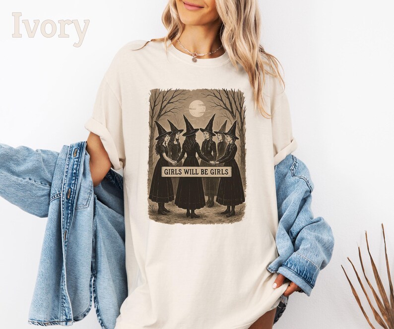 K&ouml;nnte beinhalten: Elfenbeinfarbenes T-Shirt mit einem sepiafarbenen Aufdruck von f&uuml;nf Hexen in schwarzen Kleidern und spitzen H&uuml;ten, die sich im Kreis an den H&auml;nden halten. Der Text "GIRLS WILL BE GIRLS" ist unter der Illustration zu sehen. Das Shirt hat eine lockere Passform.