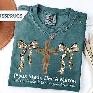May include: Teal Comfort Colors t-shirt with a cross and two bows in a camouflage pattern. The text "Jesus Made Her A Mama and she wouldn't have it any other way" is printed on the shirt. A gold necklace is draped across the shirt.