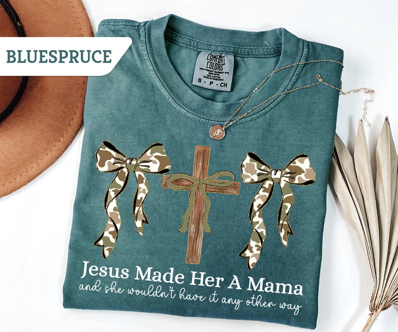 May include: Teal Comfort Colors t-shirt with a cross and two bows in a camouflage pattern. The text "Jesus Made Her A Mama and she wouldn't have it any other way" is printed on the shirt. A gold necklace is draped across the shirt.