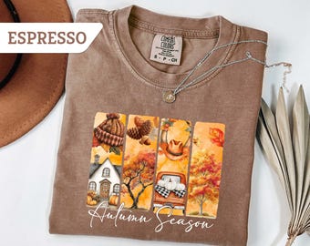 May include: Espresso-colored t-shirt with an autumn-themed graphic. The graphic features a house, a truck with pumpkins, and fall foliage. The words "Autumn Season" are written in cursive. A silver necklace and a hat are also visible.