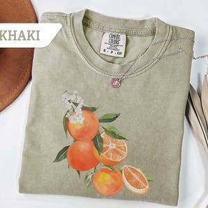 May include: Khaki-colored t-shirt featuring a watercolor illustration of oranges with green leaves and white flowers. A silver necklace with a pendant rests on the shirt. The shirt is labeled "Comfort Colors".