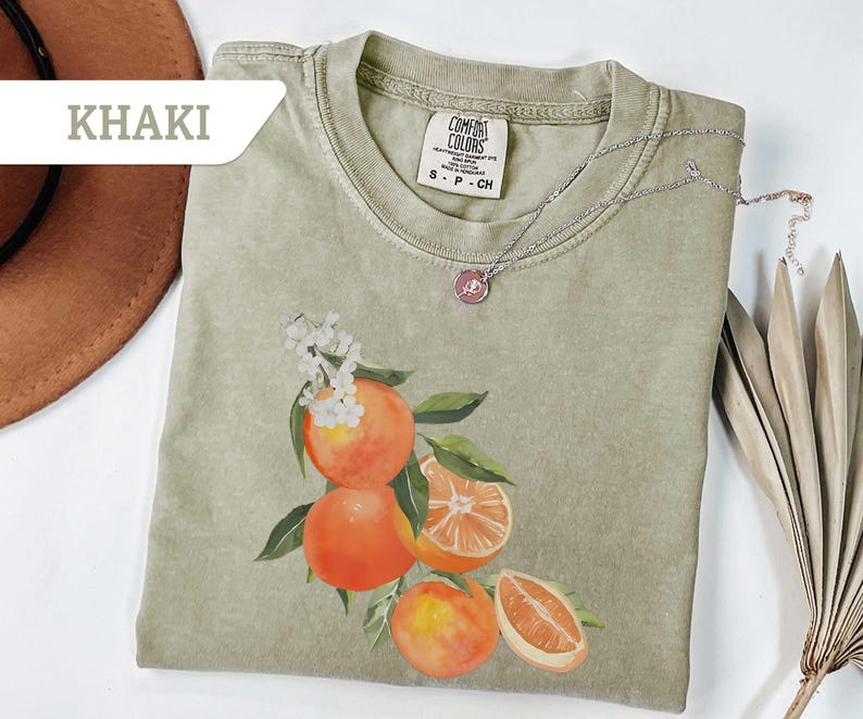 May include: Khaki-colored t-shirt featuring a watercolor illustration of oranges with green leaves and white flowers. A silver necklace with a pendant rests on the shirt. The shirt is labeled "Comfort Colors".