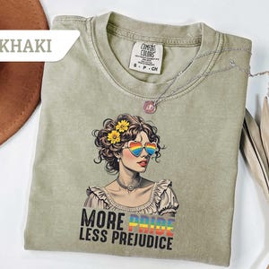 May include: Khaki-colored t-shirt featuring a graphic of a woman with rainbow heart-shaped sunglasses and yellow flowers in her hair. The text "MORE PRIDE LESS PREJUDICE" is printed below the graphic. A silver necklace is draped across the shirt.