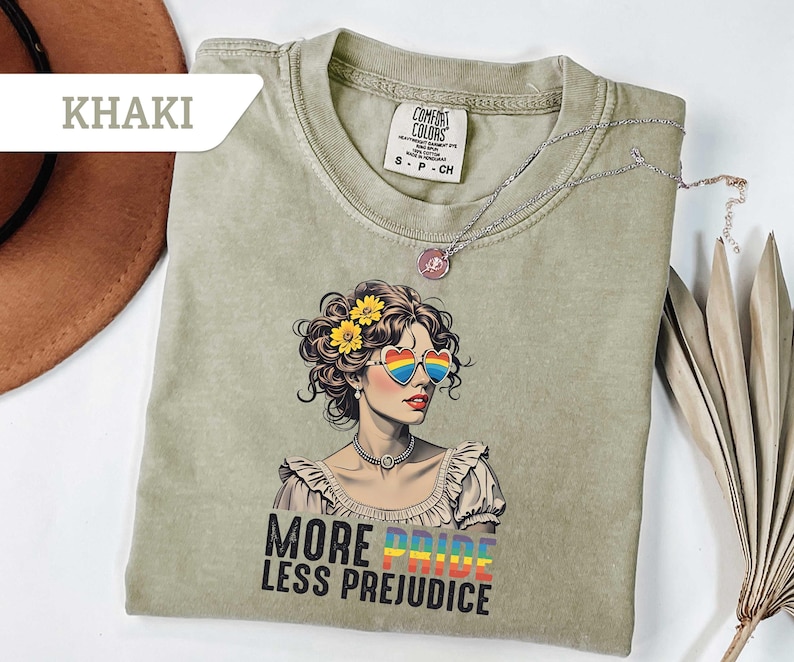 May include: Khaki-colored t-shirt featuring a graphic of a woman with rainbow heart-shaped sunglasses and yellow flowers in her hair. The text "MORE PRIDE LESS PREJUDICE" is printed below the graphic. A silver necklace is draped across the shirt.